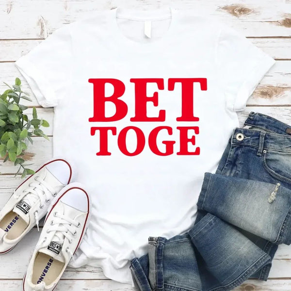 White t-shirt with 'BET TOGE' text, blue jeans, white sneakers, and a plant on a wooden surface.