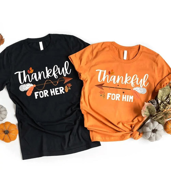 May include two matching Thanksgiving-themed T-shirts laid flat, one black reading “Thankful For Her” and one orange reading “Thankful For Him.” Both designs feature autumn leaves, feathers, and an arrow motif, surrounded by small pumpkins and fall decor.
