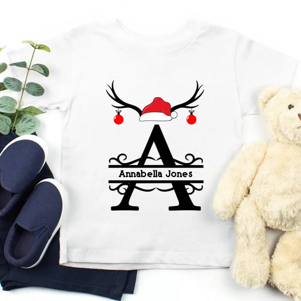 May include a festive Christmas t-shirt featuring a large custom initial printed in a holiday-themed design, laid flat with seasonal decorations.
