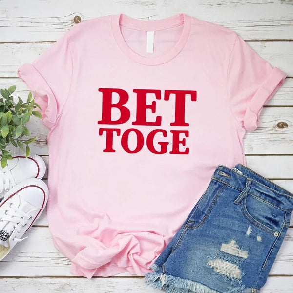 Pink t-shirt with 'BET TOGE' text, paired with denim shorts and sneakers on a wooden surface.