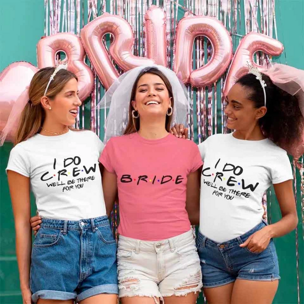 Three women wearing white 'I DO CREW' and pink 'BRIDE' t-shirts, standing in front of a 'Bride' balloon backdrop.