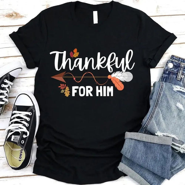 May include two matching Thanksgiving-themed T-shirts laid flat, one black reading “Thankful For Her” and one orange reading “Thankful For Him.” Both designs feature autumn leaves, feathers, and an arrow motif, surrounded by small pumpkins and fall decor.