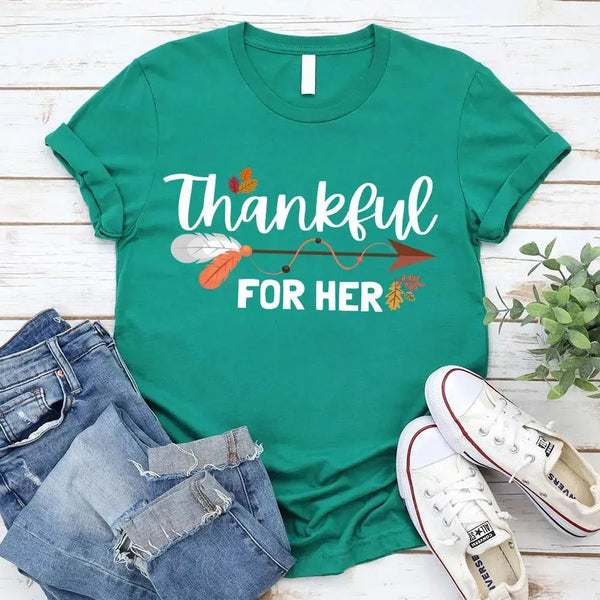 May include two matching Thanksgiving-themed T-shirts laid flat, one black reading “Thankful For Her” and one orange reading “Thankful For Him.” Both designs feature autumn leaves, feathers, and an arrow motif, surrounded by small pumpkins and fall decor.