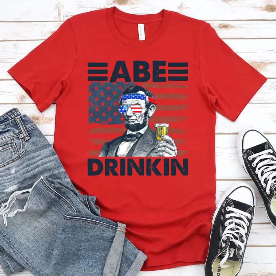 Red t-shirt with graphic of a historical figure drinking, paired with jeans and sneakers on a wooden surface.