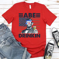 Red t-shirt with graphic of a historical figure drinking, paired with jeans and sneakers on a wooden surface.