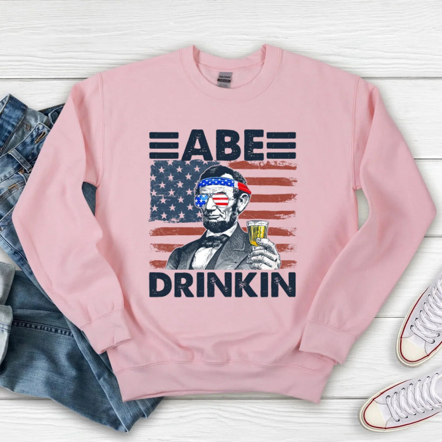 Pink sweatshirt with graphic design of a man holding a drink and American flag, text 'ABE DRINKIN', on a wooden surface with jeans and sneakers.