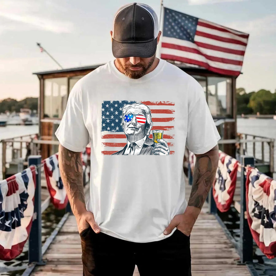 Man wearing a white t-shirt with a graphic design of a person holding a drink, with an American flag in the background, standing on a dock with American flags.