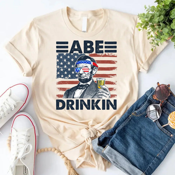 Beige custom tee with Abraham Lincoln graphic, beer, and USA flag, styled with denim shorts.
