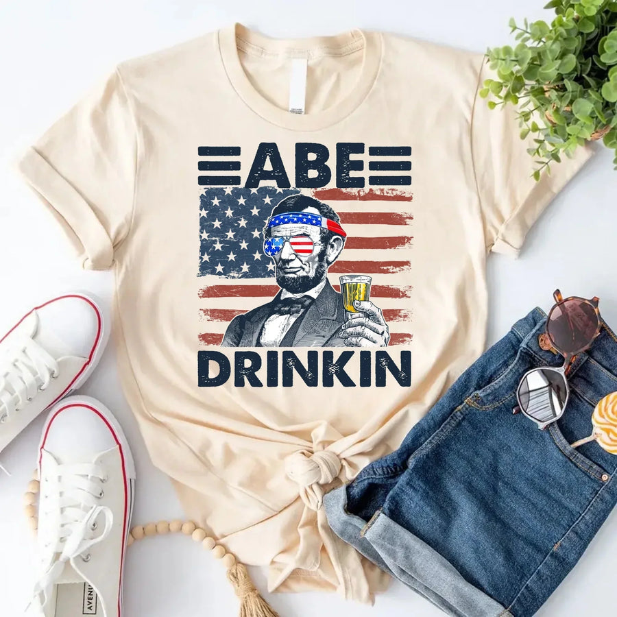 Beige custom tee with Abraham Lincoln graphic, beer, and USA flag, styled with denim shorts.