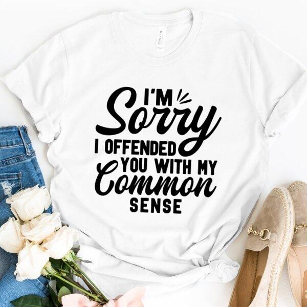 White custom tee with black text saying 'I'm sorry I offended you with my common sense', styled flatlay with jeans, flowers, and shoes