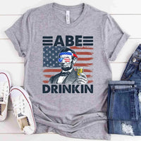 Gray t-shirt with graphic design of a historical figure holding a drink, American flag, and text 'ABE DRINKIN'.