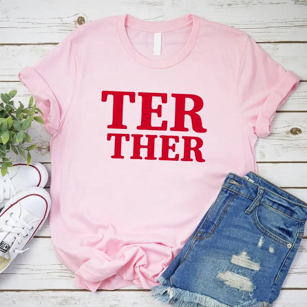 Pink t-shirt with red text 'TER THER', white sneakers, and denim shorts on a wooden surface.