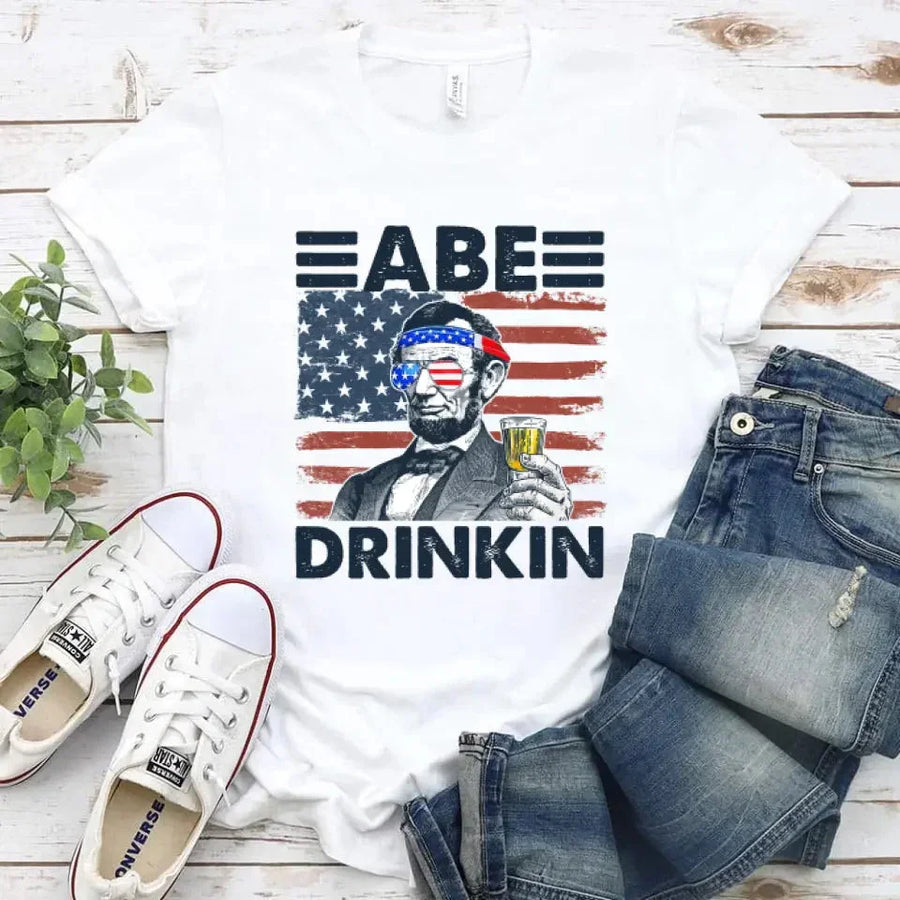 White t-shirt with 'ABE DRINKIN' graphic, jeans, and sneakers on a wooden surface.