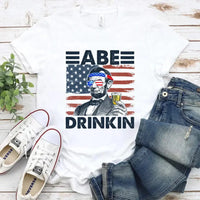 White t-shirt with 'ABE DRINKIN' graphic, jeans, and sneakers on a wooden surface.