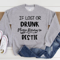 Gray sweatshirt with text 'If lost or drunk, please return to bestie' on a wooden surface with jeans and shoes.