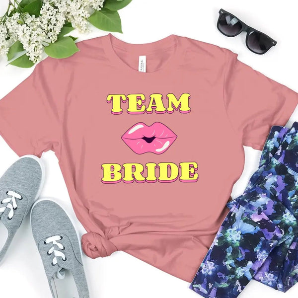 A dusty-rose sleeveless racerback shirt with graphic text “TEAM BRIDE” and a pink lips design, styled in a flat lay with sneakers, sunglasses, and flowers.