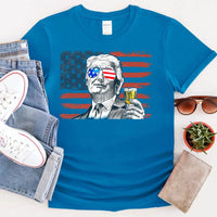Blue t-shirt with a graphic design of a person holding a drink, surrounded by jeans, sneakers, sunglasses, and a plant.