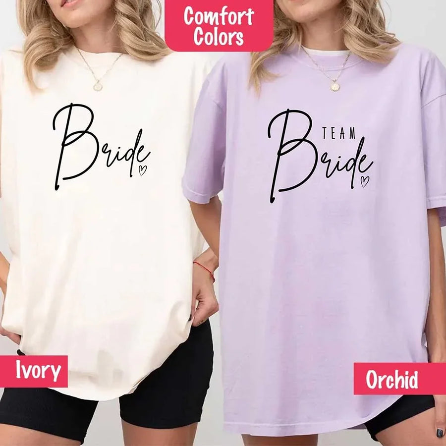 Two women wearing Comfort Colors shirts, one in Ivory with black “Bride” script, the other in Orchid purple with black “Team Bride” text and a heart accent.