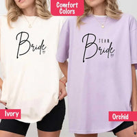 Two women wearing Comfort Colors shirts, one in Ivory with black “Bride” script, the other in Orchid purple with black “Team Bride” text and a heart accent.