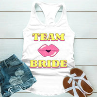 A white tank top with 'TEAM BRIDE' text and a heart symbol replacing the word 'TEAM'. The tank top is displayed alongside denim shorts and white sandals, suggesting a casual outfit for a bachelorette party.