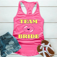 A Pink tank top with 'TEAM BRIDE' text and a heart symbol replacing the word 'TEAM'. The tank top is displayed alongside denim shorts and white sandals, suggesting a casual outfit for a bachelorette party.