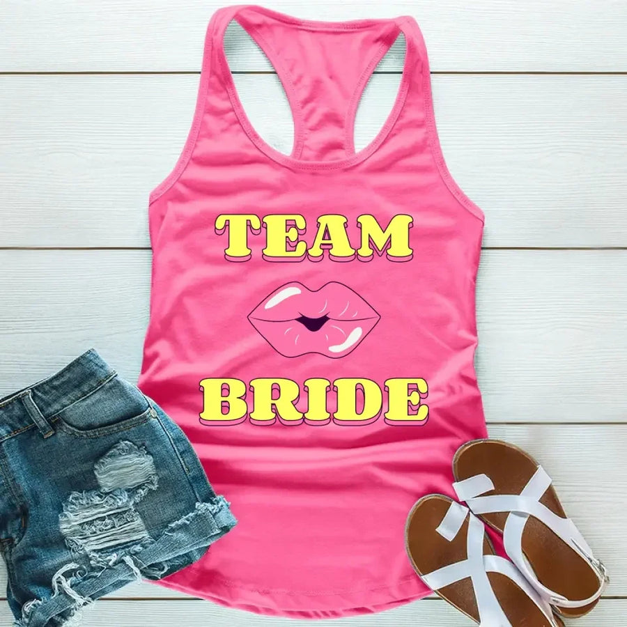 A Pink tank top with 'TEAM BRIDE' text and a heart symbol replacing the word 'TEAM'. The tank top is displayed alongside denim shorts and white sandals, suggesting a casual outfit for a bachelorette party.