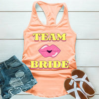 A Peach tank top with 'TEAM BRIDE' text and a heart symbol replacing the word 'TEAM'. The tank top is displayed alongside denim shorts and white sandals, suggesting a casual outfit for a bachelorette party.
