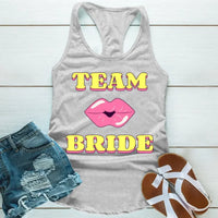 A Grey tank top with 'TEAM BRIDE' text and a heart symbol replacing the word 'TEAM'. The tank top is displayed alongside denim shorts and white sandals, suggesting a casual outfit for a bachelorette party.