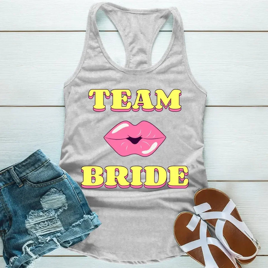 A Grey tank top with 'TEAM BRIDE' text and a heart symbol replacing the word 'TEAM'. The tank top is displayed alongside denim shorts and white sandals, suggesting a casual outfit for a bachelorette party.