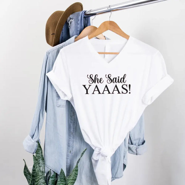 White t-shirt with 'She Said YAAAS!' text on a rack with denim jacket and hat.